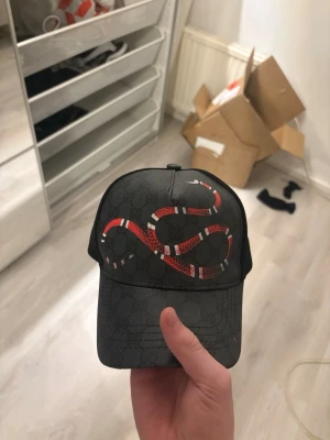 Svart keps - Svart premiumkeps med diskret monogrammönster och iögonfallande röd orm-design på framsidan. Modern trucker-modell med mesh baktill för skön passform och ventilation. Passar perfekt till både vardag och streetwear-stil.