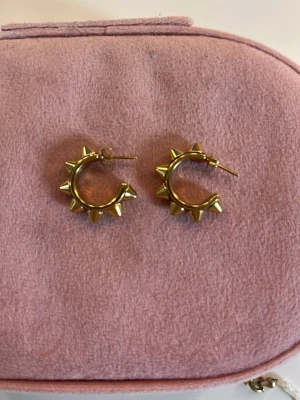 Guldiga hoops med nitar - Coola örhängen i form av små hoops i guldfärgad metall med spetsiga nitar runt hela ringen. Perfekta för dig som vill ha en edgy och trendig stil. Säljs i par.