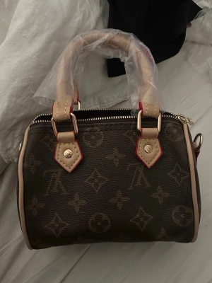 monogram handväska - Snygg handväska i klassiskt brunt monogrammönster med beige detaljer och guldfärgade metalldelar. Väskan har både handtag och avtagbar axelrem, perfekt för dig som gillar lyxig och tidlös design. Materialet är canvas med läderdetaljer.