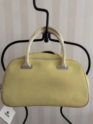 Gul handväska från Lacoste - Snygg gul handväska från Lacoste i canvas med två korta handtag och diskret krokodil-logga framtill. Väskan har en mjuk, rundad form och dragkedja upptill. Perfekt för dig som vill ha en stilren och trendig accessoar.