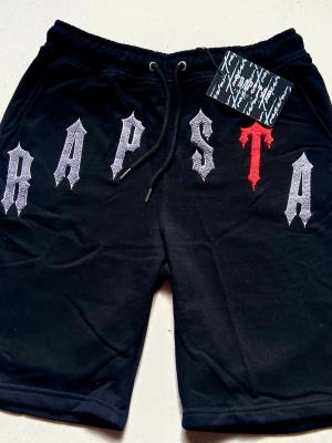 Svart Trapstar t-shirt och shorts bundle  - Svart t-shirt från Trapstar med stort tryck i vitt och rött på bröstet där det står 'TRAPSTAR' i gotisk stil. T-shirten har rund hals, korta ärmar och är gjord i mjuk bomull. Perfekt för en streetwear-look.