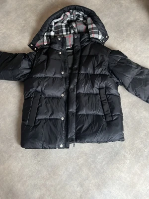 Svart dunjacka med avtagbar huva - Säljer en svart dunjacka med avtagbar huva och dragkedja. Jackan har tryckknappar och är tillverkad i ett glansigt material som ger en modern look. Perfekt för kalla vinterdagar och riktigt varm.