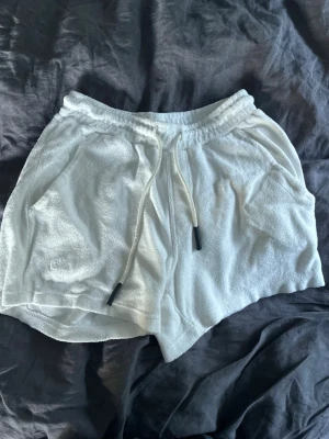 Vita frottéshorts  OAS - Säljer ett par vita shorts i mjuk frotté med elastisk midja och snörning framtill från OAS. Shortsen har två sidofickor och en bakficka, perfekt för en chill sommarlook. Lätta och sköna att bära, passar perfekt till varma dagar.