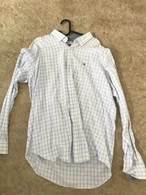 Vit rutig skjorta från Tommy Hilfiger - Vit skjorta från Tommy Hilfiger med blått rutmönster och klassisk krage. Skjortan har långa ärmar, knappar framtill och en liten logga på bröstet. Perfekt för en stilren look.
