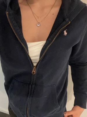 Mörkblå hoodie från Ralph Lauren - Mörkblå hoodie med dragkedja från Ralph Lauren. Klassisk design med huva, fickor framtill och den ikoniska broderade loggan på bröstet. Mjuk bomullskänsla och perfekt för en avslappnad stil.