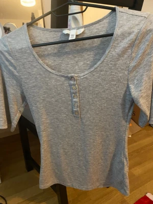 Grå ribbad topp med knappar H&M - Grå ribbad topp från H&M med rund halsringning och knappdetalj framtill. Toppen har korta ärmar och är tillverkad i ett mjukt, stretchigt material som sitter skönt på kroppen. Perfekt att styla med jeans eller kjol.