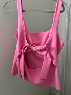 Rosa linnetopp med rosett - Supersöt rosa linnetopp med breda axelband och en stor rosett baktill. Toppen har en fyrkantig urringning och är figurnära i modellen. Perfekt för dig som vill sticka ut med en färgglad och trendig look. Kan bära denna även om du skulle vara en L!