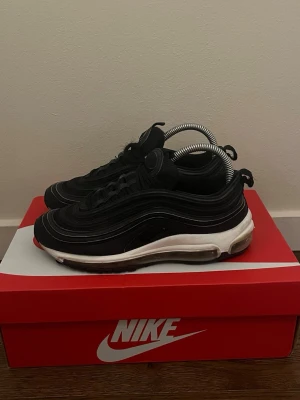 Nike Air Max 97 svarta sneakers - Nike Air Max 97 sneakers i svart med vita detaljer och synlig Air Max-sula. Skorna har en vågformad design, reflekterande detaljer och snörning framtill. Ovandelen är i textil och syntet, sulan är i gummi. Perfekta för en streetwear-look.