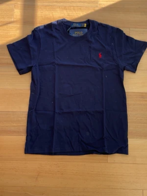 Marinblå Ralph Lauren T-whirt - 👕 Märke: Polo Ralph Lauren 📏 Storlek: M 🎨 Färg: Marinblå med röd logga ✨ ☀️ Stilren och snygg t-shirt från Polo Ralph Lauren – perfekt för sommaren och enkel att matcha med både shorts och jeans. Tillverkad i mjuk och bekväm bomull med klassisk passform och ikonisk röd logga på bröstet.  ✨ Skick: Helt ny med tags kvar 🧼 Aldrig använd 👌 Tidlös modell som passar till allt