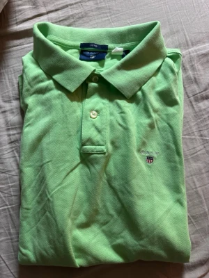 Grön pikétröja från Gant - Snygg ljusgrön pikétröja från Gant med klassisk krage och två knappar framtill. Tröjan har korta ärmar och en liten broderad logga på bröstet. Perfekt för en avslappnad och stilren look.