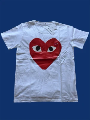 CDG tröja  - Vit t-shirt från Comme des Garçons Play med det ikoniska röda hjärtat med ögon tryckt på bröstet. Klassisk rund hals och korta ärmar. Tillverkad i mjuk bomull, perfekt för en stilren och trendig look.