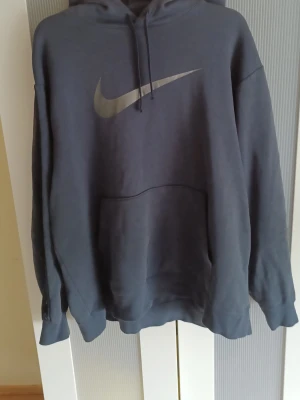 Mörkgrå Nike hoodie med logga - Snygg mörkgrå hoodie från Nike med stor silvrig swoosh-logga på bröstet och mindre loggor längs ärmarna. Klassisk känguruficka framtill och justerbar huva med dragsko. Perfekt för en sportig och avslappnad stil.