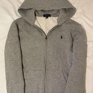 Grå hoodie från Ralph Lauren - Säljer en stilren grå hoodie från Ralph Lauren med dragkedja och huva. Perfekt för en avslappnad look. Den har en liten broderad logga på bröstet och är tillverkad i mjukt material för extra komfort. Använd Max 3ggr. Se att den är äkta genom att scanna Qr-koden på 3:dje bilden💗