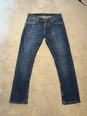 Nudie Grim Tim - Snygga blå jeans från Nudie Jeans Co med klassisk femficksmodell och raka ben. Jeansen har tydliga kontrastsömmar och är tillverkade i slitstarkt denimtyg. Perfekta för en avslappnad och trendig stil. Mått (+-1cm): Midja ~ 41cm Längd ~ 104cm Benöppning ~ 18 