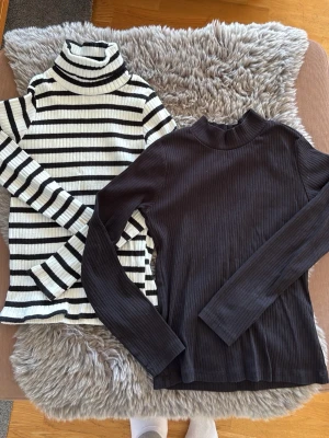 Randig och svart ribbad polotröja - Två ribbade polotröjor, en svart och en vit med svarta ränder. Båda har lång ärm och hög krage, perfekt för lager-på-lager. Materialet är stretchigt och mjukt, vilket gör dem bekväma och snygga att bära. Passar dig som gillar stilrena basics.