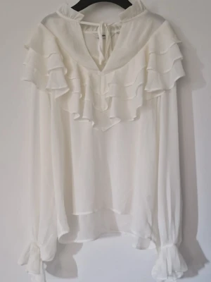 Vit volangblus med knytband - Säljer denna eleganta vita blus från Kappahl i storlek M. Blusen har fina volanger framtill och baktill med volangkant nertill. Den har även en söt knytdetalj framtill.  Jag säljer den för att den tyvärr är lite för stor för mig. Den är endast använd en gång och är i nyskick. Perfekt för en festlig tillställning eller att klä upp en vardagsoutfit!