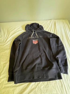 Svart hoodie från GANT med emblem - Svart hoodie från GANT med broderad logga och amerikanskt inspirerat emblem på bröstet. Hoodien har vit dragsko och stor känguruficka framtill. Perfekt för dig som gillar klassisk och sportig stil. Materialet är mjukt och bekvämt bomullstyg.
