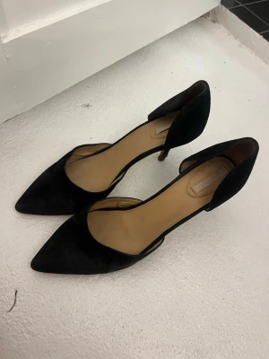Svarta klackar  - Säljer ett par eleganta svarta klackar med spetsig tå och öppen sida. Skorna har en tunn klack och är tillverkade i ett material som liknar mocka. Perfekta för dig som gillar stilrena och klassiska skor. Stolek 41