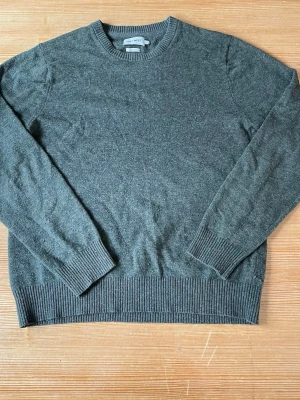 Mörkgrön stickad tröja från East west - Mysig mörkgrön stickad tröja från east west med rund halsringning och ribbade muddar vid ärmslut och nederkant. Tröjan har en enkel och stilren design, perfekt för lager på lager och passar till det mesta. Materialet är mjukt och skönt.
