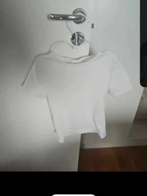 Vit croppad t-shirt i bomull - Enkel och stilren vit croppad t-shirt med korta ärmar och rund hals. Perfekt basplagg som är lätt att matcha till olika outfits. Tillverkad i mjuk bomull för en skön känsla hela dagen.