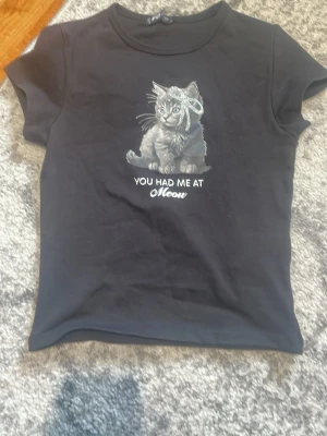 Svart t-shirt med kattmotiv - Cool svart t-shirt med ett sött kattmotiv och texten 'You had me at Meow' på framsidan. T-shirten har korta ärmar och en klassisk rund halsringning. Perfekt för dig som älskar katter och vill ha en casual look.