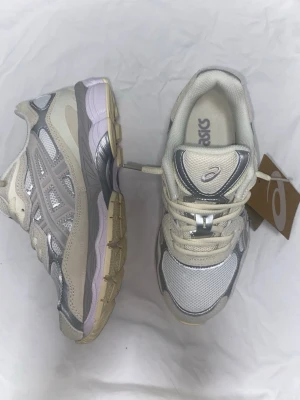 Asics Gel NYC  - Snygga Asics sneakers i beige och silver med meshpaneler och detaljer i grått. Skorna har en chunky sula i beige och lila toner samt klassisk Asics-logga på plösen. Perfekta för dig som gillar retrostil och sportig vibe.