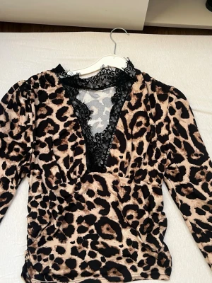 Leopardmönstrad topp med spets - Säljer en populär  leopardmönstrad långärmad topp med djup v-ringning och svart spetsdetalj runt halsen. Toppens material känns mjukt och stretchigt, perfekt för dig som vill sticka ut. Passformen är normal och toppen har coola detaljer som gör den unik.