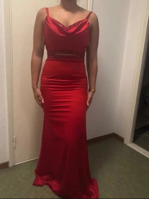 Röd långklänning med smala axelband - Elegant röd långklänning i satin med smala axelband och draperad urringning. Klänningen har markerad midja med dekorativa detaljer och figurnära passform som framhäver siluetten. Perfekt för speciella tillfällen. Aldrig använd så nytt skick. Den är köpt för 3000 kr. Jag skulle säga att de är en storlek S, då jag har de och den sitter perfekt på mig.