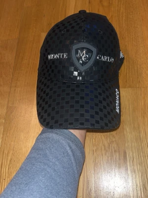 Svart keps Monte Carlo Monaco - Snygg svart keps med rutigt mönster och broderade detaljer. Framtill står det 'Monte Carlo' och ett MC-emblem, på sidan finns texten 'Monaco' och en broderad flagga med målflagg. Justerbar baktill och tillverkad i syntetmaterial. Köpt i Monaco.