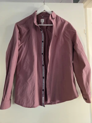 Rosa skjorta från Morris - Säljer en stilren rosa skjorta från Morris med broderad logga på bröstet. Skjortan har klassisk krage, långa ärmar och knappar framtill. Tillverkad i mjuk bomull som känns skön mot huden. Perfekt för dig som vill ha en enkel men snygg skjorta.