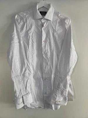 Vit slim fit skjorta från Dressmann - Klassisk vit skjorta från Dressmann i slim fit-modell. Tillverkad i stretchig bomull för extra komfort och har knappar framtill samt vid ärmsluten. Perfekt för en clean och stilren look.