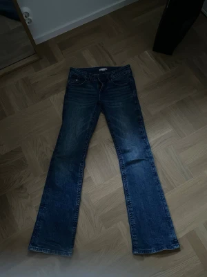 Bootcut jeans med broderade fickor - Säljer ett par mörkblå bootcut jeans med snygga broderade detaljer och knappar på bakfickorna. Klassisk femficksmodell med låg midja och slitningar framtill. Perfekt för dig som gillar en avslappnad och trendig look.