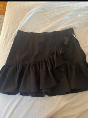 Svart volangkjol  - Säljer en svart kortkjol med volangdetaljer och omlottkänsla. Kjolen har en snygg och trendig look som passar perfekt till många olika outfits. Tillverkad i ett mjukt och lätt material som ger en skön känsla. Helt ny oanvänd 
