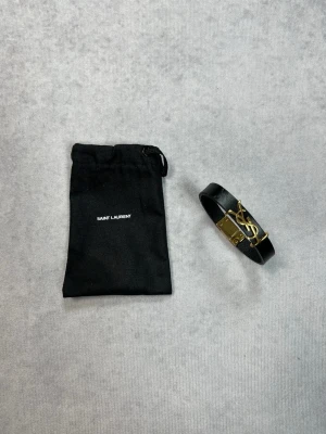 Saint Laurent svart läderarmband - Stilrent svart armband i läder från Saint Laurent med ikoniskt YSL-logo i guldmetall på framsidan. Armbandet har en elegant och tunn design, och kommer med en svart tygpåse med Saint Laurent-tryck. Perfekt för dig som gillar exklusiva accessoarer.