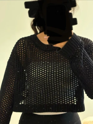 Svart stickad tröja croped - Säljer en svart stickad croptröja i meshstil med grova hål och rund hals. Tröjan har långa ärmar och ribbade muddar. Perfekt för lager på lager och ger en cool, edgy vibe till outfiten.