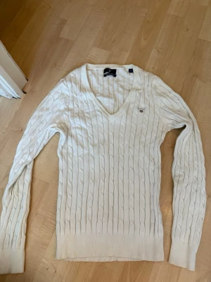 Vit kabelstickad tröja från Gant - Snygg vit kabelstickad tröja från Gant med V-ringning och diskret logga på bröstet. Tröjan har långa ärmar och ribbade muddar vid ärmslut och nederkant. Perfekt till jeans eller kjol för en clean och klassisk look.
