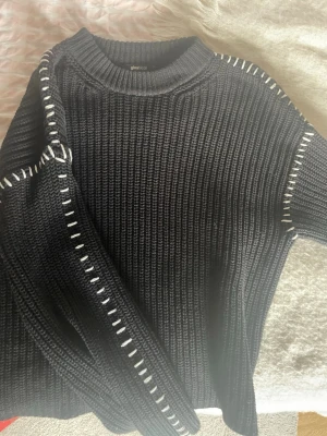 Svart stickad tröja från Gina Tricot - Cool svart stickad tröja från Gina Tricot med vita dekorativa sömmar längs axlar och ärmar. Tröjan har rund halsringning och är ribbstickad för en snygg struktur. Perfekt för dig som gillar stilrena detaljer och enkel design.