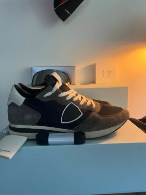 Grå och blå sneakers från Philippe Model - Snygga sneakers från Philippe Model med grå mockadetaljer, mörkblå textil och vita skosnören. Skorna har en låg profil och en vit kant runt sulan. Ikonisk logotyp på sidan och bekväm passform. Perfekta för dig som gillar stilrena och trendiga sneakers.