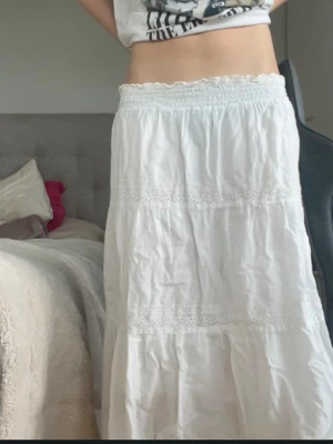 Vit långkjol med spetsdetaljer - Säljer en vit långkjol med elastisk midja och fina spetsdetaljer. Kjolen är luftig och har ett romantiskt uttryck med flera lager och broderade band. Perfekt för dig som gillar en bohemisk och avslappnad stil.