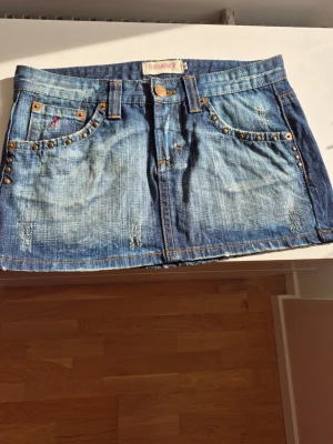 Jeanskjol  - Snygg blå kort jeanskjol från Terranova med slitningar och coola nitar på fickorna både fram och bak. Klassisk femficksmodell med knapp och dragkedja. Perfekt för dig som gillar en edgy och trendig stil. Rätt kort och knappar använd. Storlek M. Längden är 27 cm och höftmåt 80 cm.