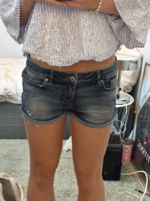 Blå jeansshorts från LTB - Säljer ett par blå denimshorts från LTB med klassisk femficksdesign och slitna detaljer. Shortsen har låg midja, uppvikta benslut och en snygg tvättad look. Perfekta till sommaren och lätta att matcha med olika toppar.