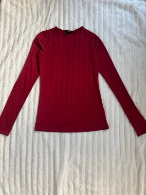 Röd långärmad topp från Lindex - En stilren vinröd långärmad topp från Lindex med rund halsringning. Modellen är figurnära och enkel utan mönster eller detaljer, perfekt att matcha med olika outfits. Materialet känns mjukt och stretchigt, vilket gör den bekväm att bära hela dagen.