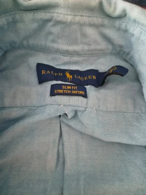 Blå slim fit skjorta från Ralph Lauren - Säljer en ljusblå slim fit stretch oxford-skjorta från Ralph Lauren med klassisk krage och knappar framtill. Skjortan har lång ärm och ikonisk broderad logga på bröstet. Perfekt för dig som gillar stilrena och tidlösa plagg.