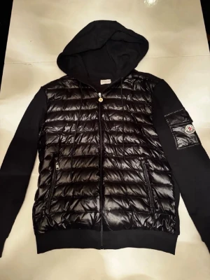 Svart quiltad jacka med huva Moncler - Svart jacka från Moncler med quiltad, glansig front i polyester och mjuka ärmar samt huva i bomull. Jackan har dragkedja framtill, fickor med dragkedja och Moncler-logga på ärmen. Perfekt för dig som gillar en sportig och modern stil. Pris kan diskuteras, öppen för byten, kontakta vid intresse.