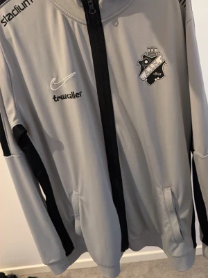 Grå AIK Nike träningsjacka - Snygg grå träningsjacka från Nike med AIK:s klubbmärke på bröstet och svarta detaljer längs ärmarna. Jackan har hel dragkedja, fickor och tryckta sponsorer som Truecaller, Fabege och SVEA. Perfekt för träning eller att bära till vardags.