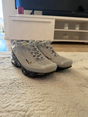 Nike air Max plus  - Grå Nike Tuned Air sneakers med svarta och gula detaljer. Skorna har en futuristisk design med synlig dämpning i sulan, snörning och små gula swoosh-loggor på sidan. Ovandelen är i syntetmaterial och mesh för bra andningsförmåga. Storlek 40 men känns som 41