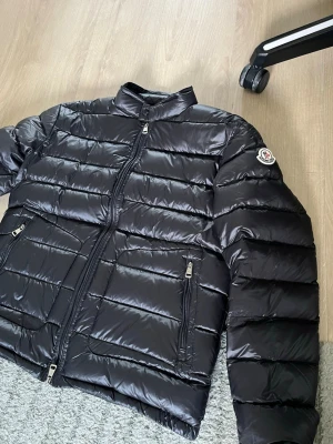 Svart dunjacka från Moncler - Säljer en svart dunjacka från Moncler med quiltad design och hög krage. Jackan har två fickor med dragkedja och Moncler-logga på ena ärmen. Perfekt för kalla vinterdagar och riktigt snygg att bära till jeans eller joggers.