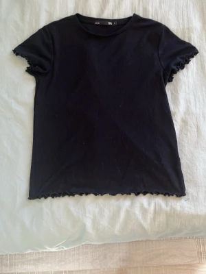 Svart ribbad t-shirt med volangkant - Säljer en svart ribbad t-shirt från Lager 157 med korta ärmar och volangkant vid ärmslut och nederkant. T-shirten har en klassisk rund halsringning och är gjord i mjuk bomullsmix. Perfekt till jeans eller kjol för en enkel men snygg look.