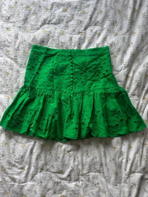 Grön volangkjol från Zara - Säljer en grön kortkjol från Zara med volangkant och broderade hål. Kjolen har en figurnära passform upptill och en utställd nederdel 