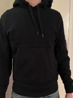 Svart hoodie från C.P. Company - Säljer en svart hoodie från C.P. Company med klassisk huva och dragsnören. Hoodien har en stor magficka och en extra ficka med dragkedja på ärmen. Tillverkad i mjukt bomullsmaterial som är skönt att bära. Perfekt för en avslappnad och stilren look.